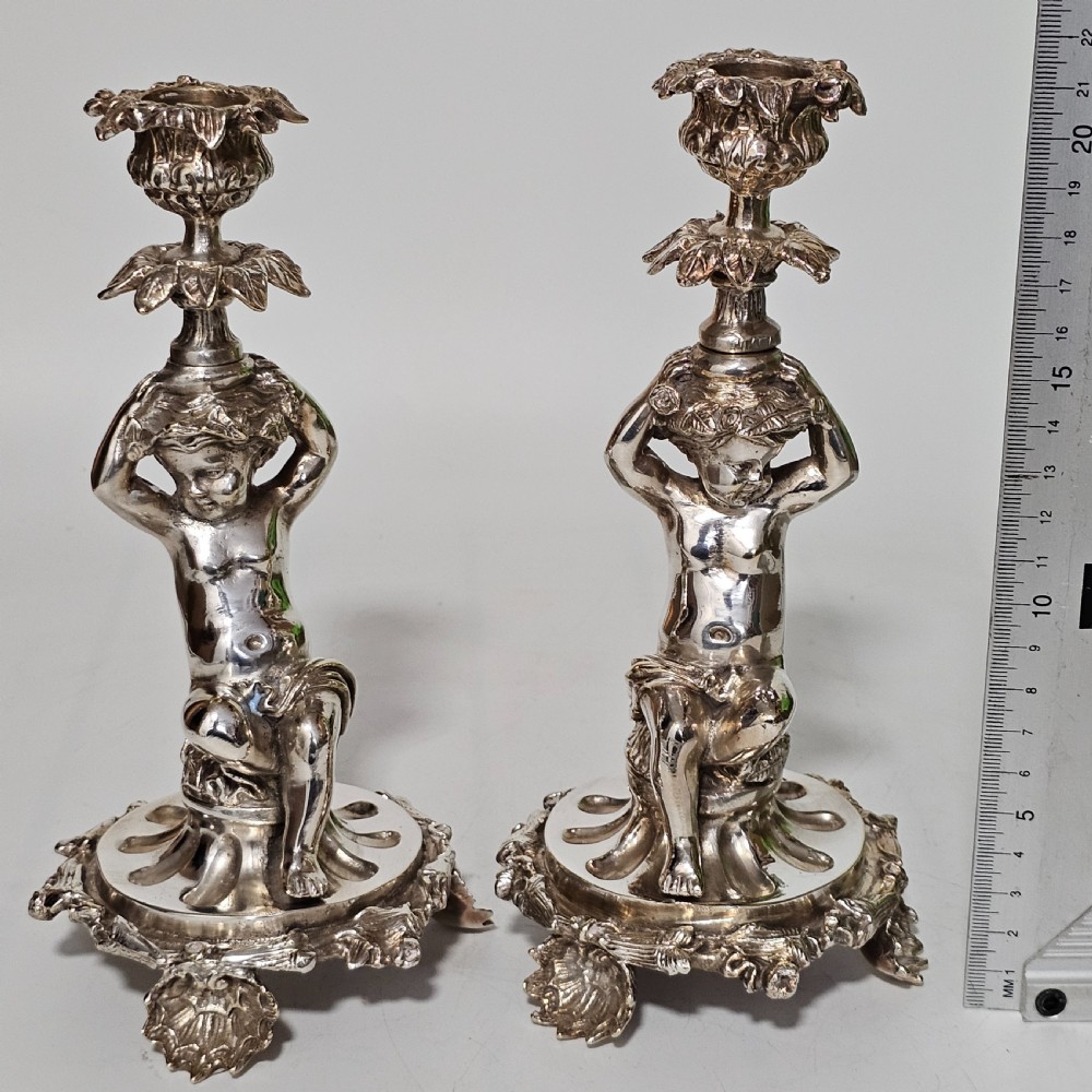 A pair of antique silver-plated candlesticks featuring seated cherub figures supporting the candle nozzles above circular scrolled bases. View XIII