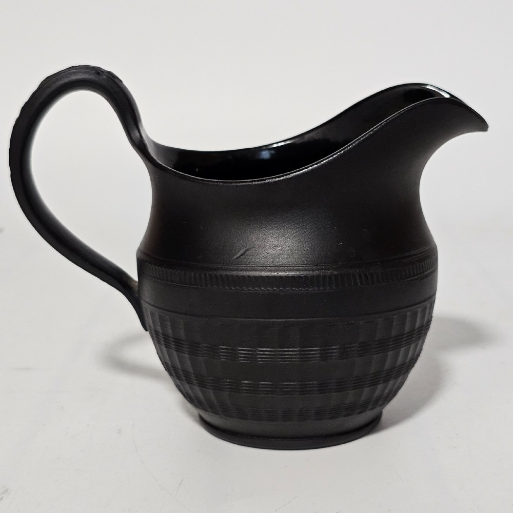 A dark matte black stoneware cream jug featuring incised geometric and reeded bands with a curved handle and pointed pouring spout. View IX