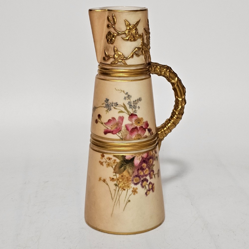 A Royal Worcester blush ivory porcelain ewer featuring hand-painted floral motifs, a gilt textured handle, and raised gold blossom decoration on the neck.