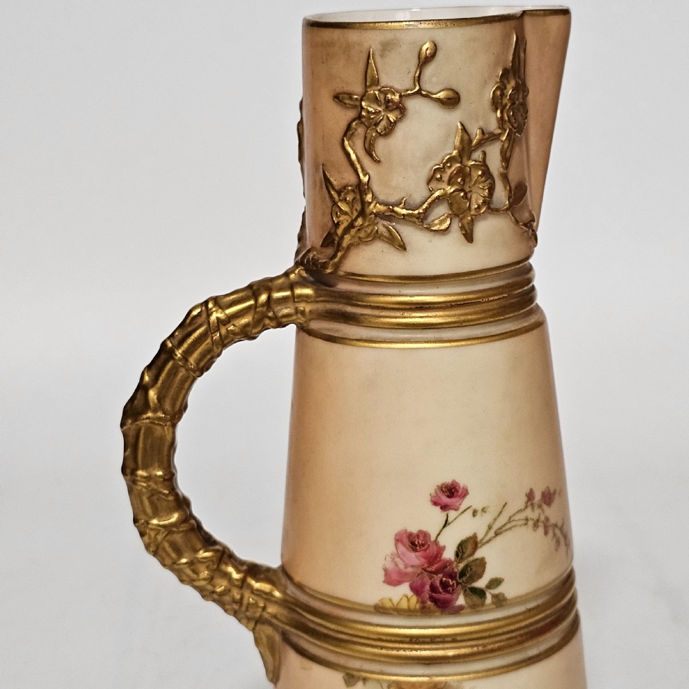 A Royal Worcester blush ivory porcelain ewer featuring hand-painted floral motifs, a gilt textured handle, and raised gold blossom decoration on the neck. View III