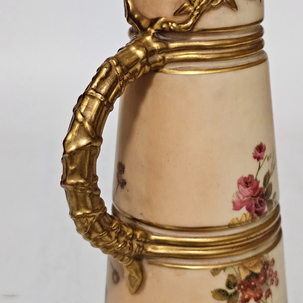 A Royal Worcester blush ivory porcelain ewer featuring hand-painted floral motifs, a gilt textured handle, and raised gold blossom decoration on the neck. View V