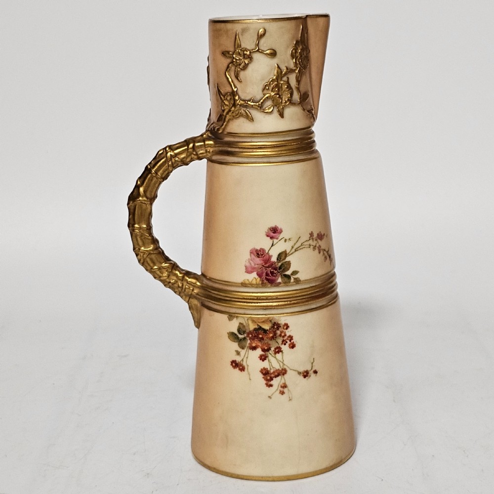 A Royal Worcester blush ivory porcelain ewer featuring hand-painted floral motifs, a gilt textured handle, and raised gold blossom decoration on the neck. View VII