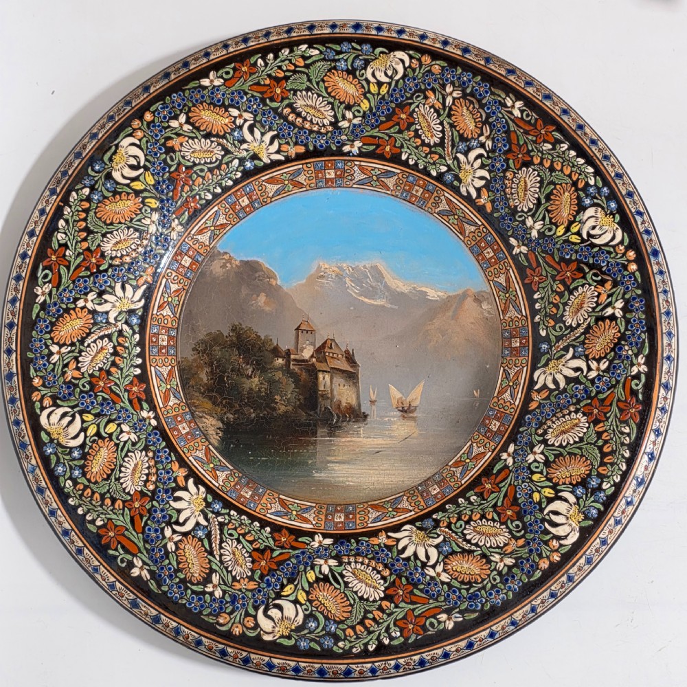 A large circular ceramic charger featuring a hand-painted central landscape of a lakeside castle with alpine mountains, surrounded by a dark border filled with dense polychrome floral motifs.