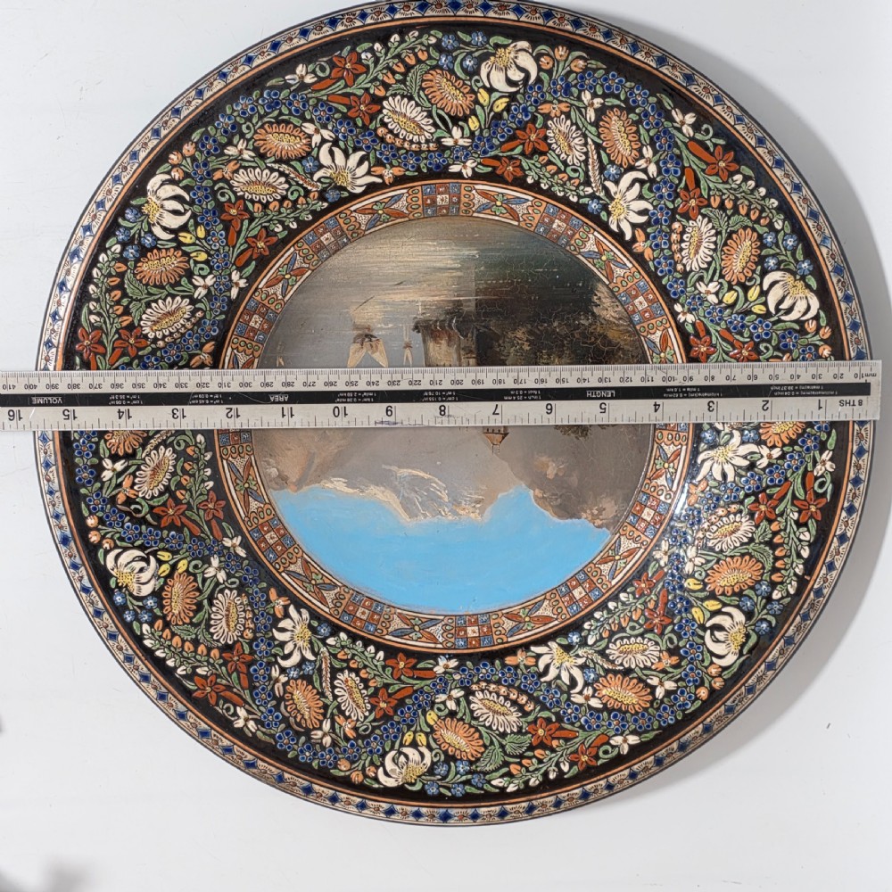 A large circular ceramic charger featuring a hand-painted central landscape of a lakeside castle with alpine mountains, surrounded by a dark border filled with dense polychrome floral motifs. View II