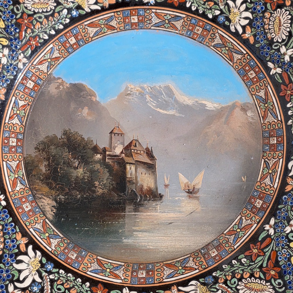 A large circular ceramic charger featuring a hand-painted central landscape of a lakeside castle with alpine mountains, surrounded by a dark border filled with dense polychrome floral motifs. View III