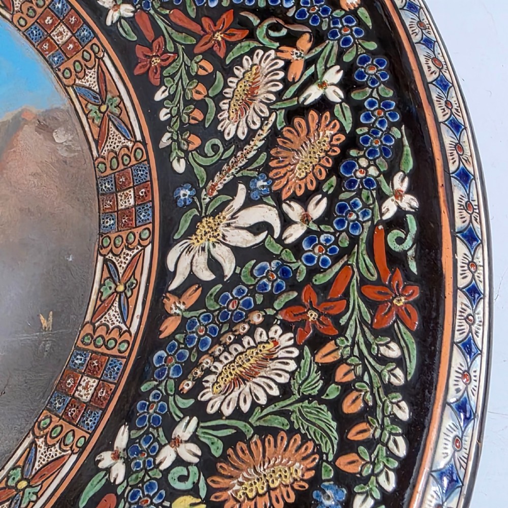 A large circular ceramic charger featuring a hand-painted central landscape of a lakeside castle with alpine mountains, surrounded by a dark border filled with dense polychrome floral motifs. View IV