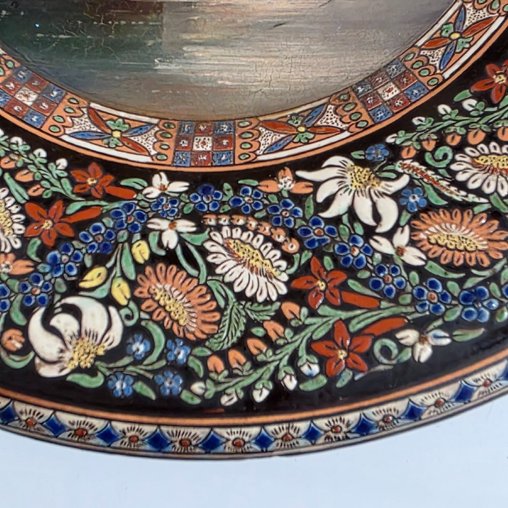 A large circular ceramic charger featuring a hand-painted central landscape of a lakeside castle with alpine mountains, surrounded by a dark border filled with dense polychrome floral motifs. View V