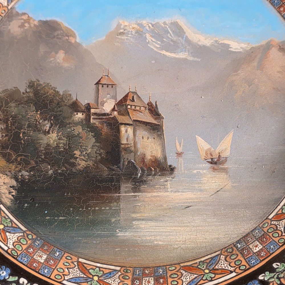 A large circular ceramic charger featuring a hand-painted central landscape of a lakeside castle with alpine mountains, surrounded by a dark border filled with dense polychrome floral motifs. View VI