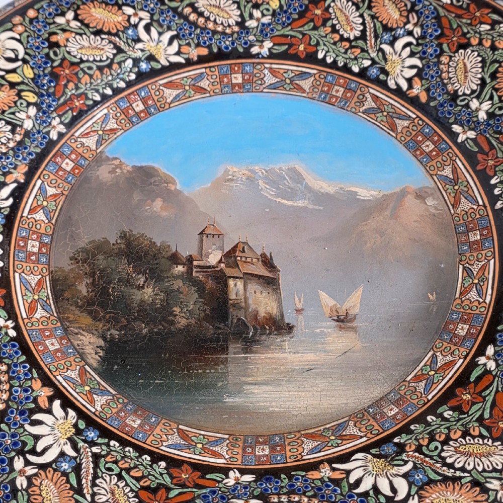 A large circular ceramic charger featuring a hand-painted central landscape of a lakeside castle with alpine mountains, surrounded by a dark border filled with dense polychrome floral motifs. View VII