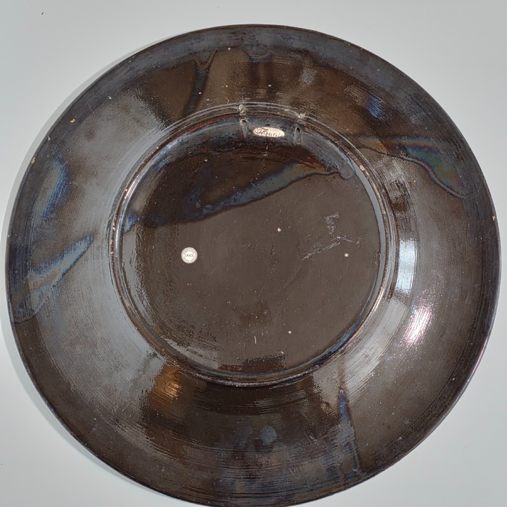 A large circular ceramic charger featuring a hand-painted central landscape of a lakeside castle with alpine mountains, surrounded by a dark border filled with dense polychrome floral motifs. View VIII