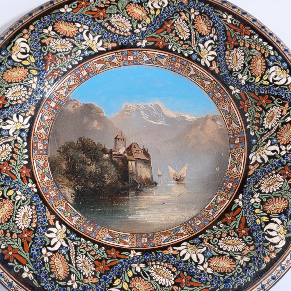 A large circular ceramic charger featuring a hand-painted central landscape of a lakeside castle with alpine mountains, surrounded by a dark border filled with dense polychrome floral motifs. View XI