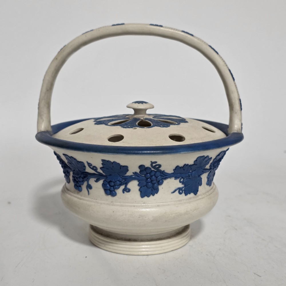 An off-white Georgian stoneware pot pourri basket featuring an overhead handle, pierced cover, and cobalt blue applied grapevine sprig decoration.