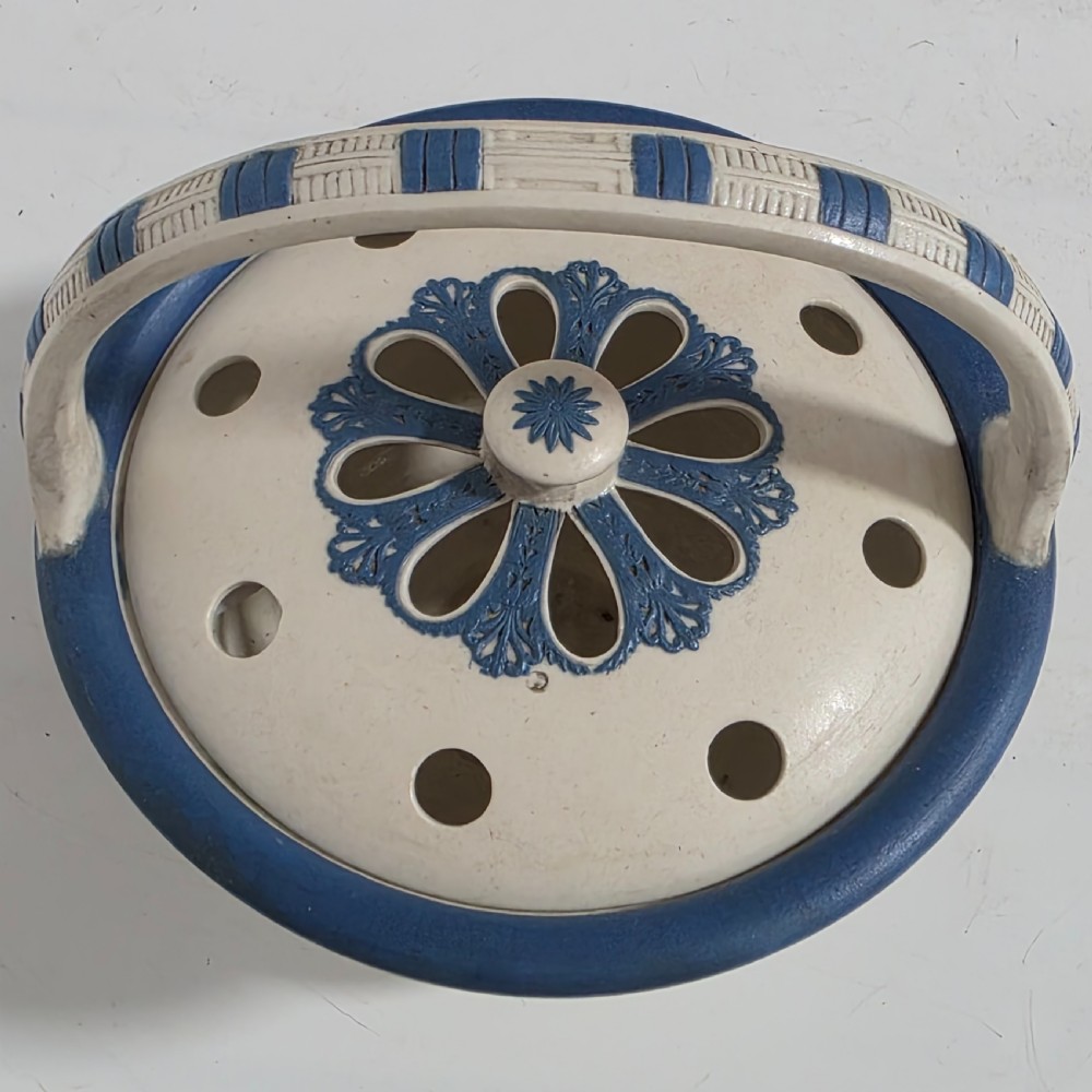 An off-white Georgian stoneware pot pourri basket featuring an overhead handle, pierced cover, and cobalt blue applied grapevine sprig decoration. View V