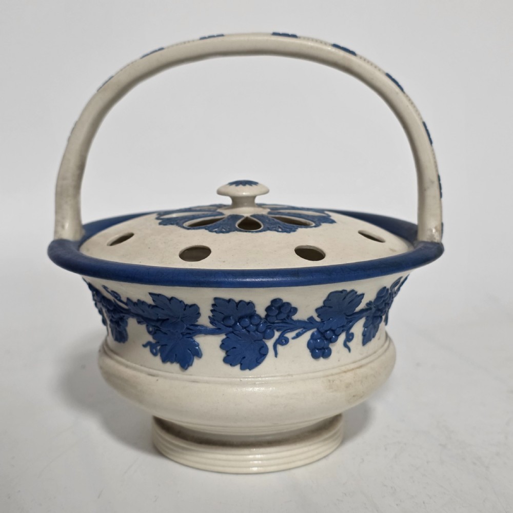 An off-white Georgian stoneware pot pourri basket featuring an overhead handle, pierced cover, and cobalt blue applied grapevine sprig decoration. View XIII
