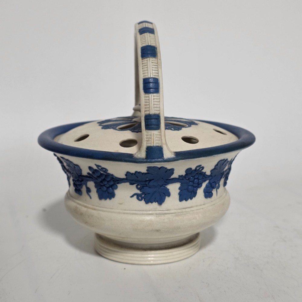 An off-white Georgian stoneware pot pourri basket featuring an overhead handle, pierced cover, and cobalt blue applied grapevine sprig decoration. View XIV