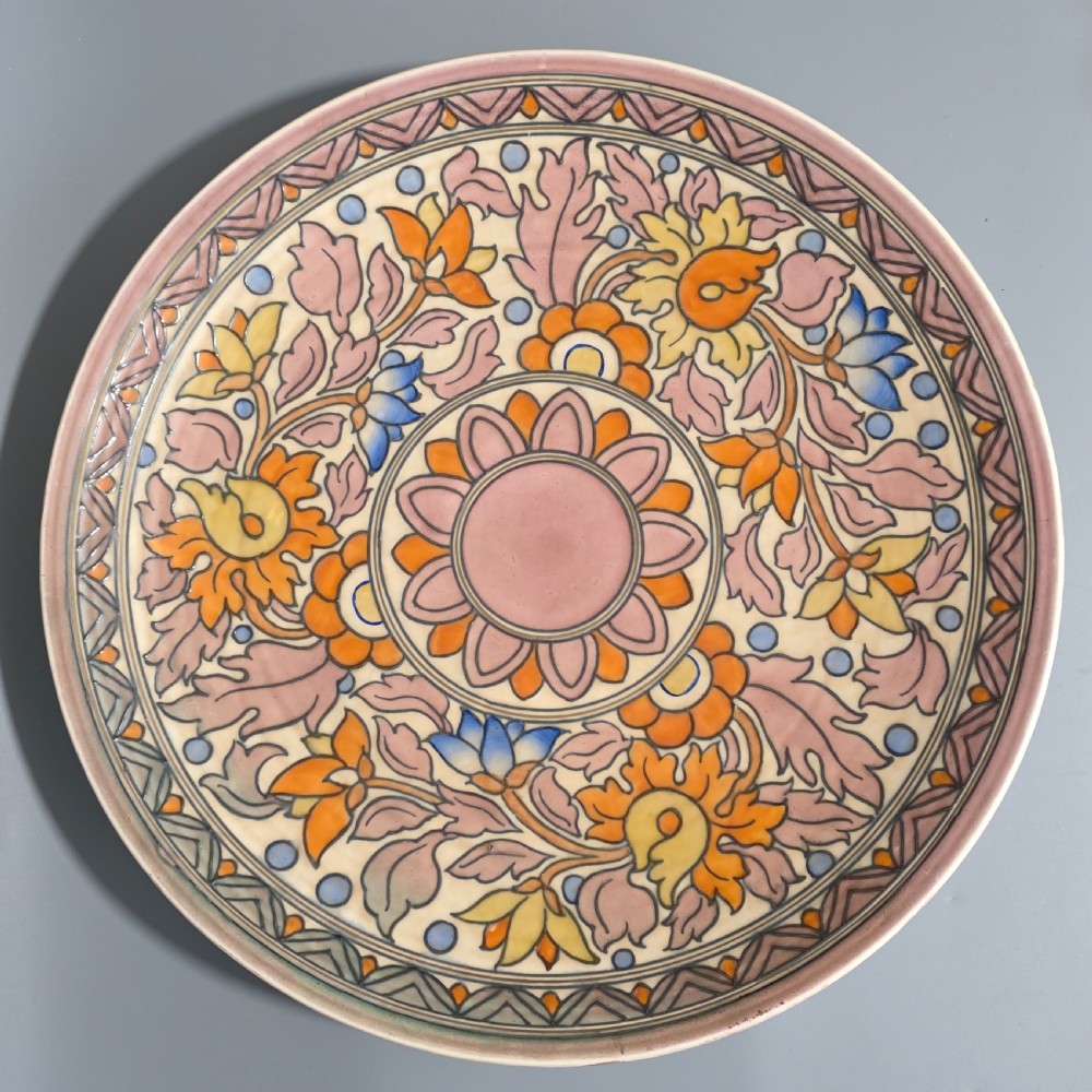 A circular Art Deco earthenware charger decorated with pink, orange, yellow, and blue stylised floral tubelined motifs.
