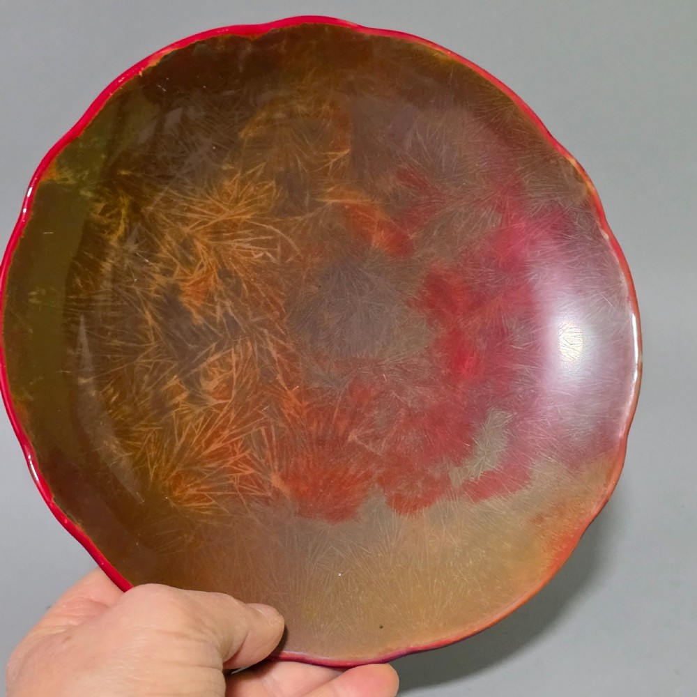 A shallow ceramic art pottery plate featuring a crimson red flambe glaze combined with golden-brown crystalline textures and a scalloped edge. View IX