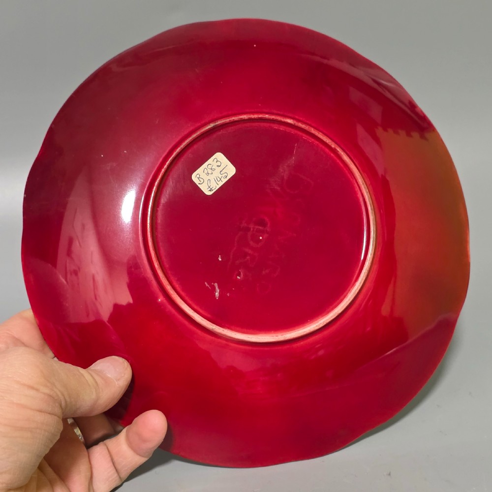 A shallow ceramic art pottery plate featuring a crimson red flambe glaze combined with golden-brown crystalline textures and a scalloped edge. View X