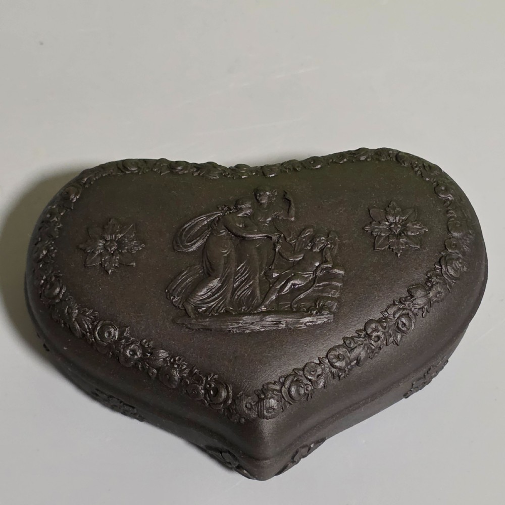 A kidney-shaped matte black basalt stoneware trinket box featuring Neoclassical relief figures, rosettes, and floral garland borders.