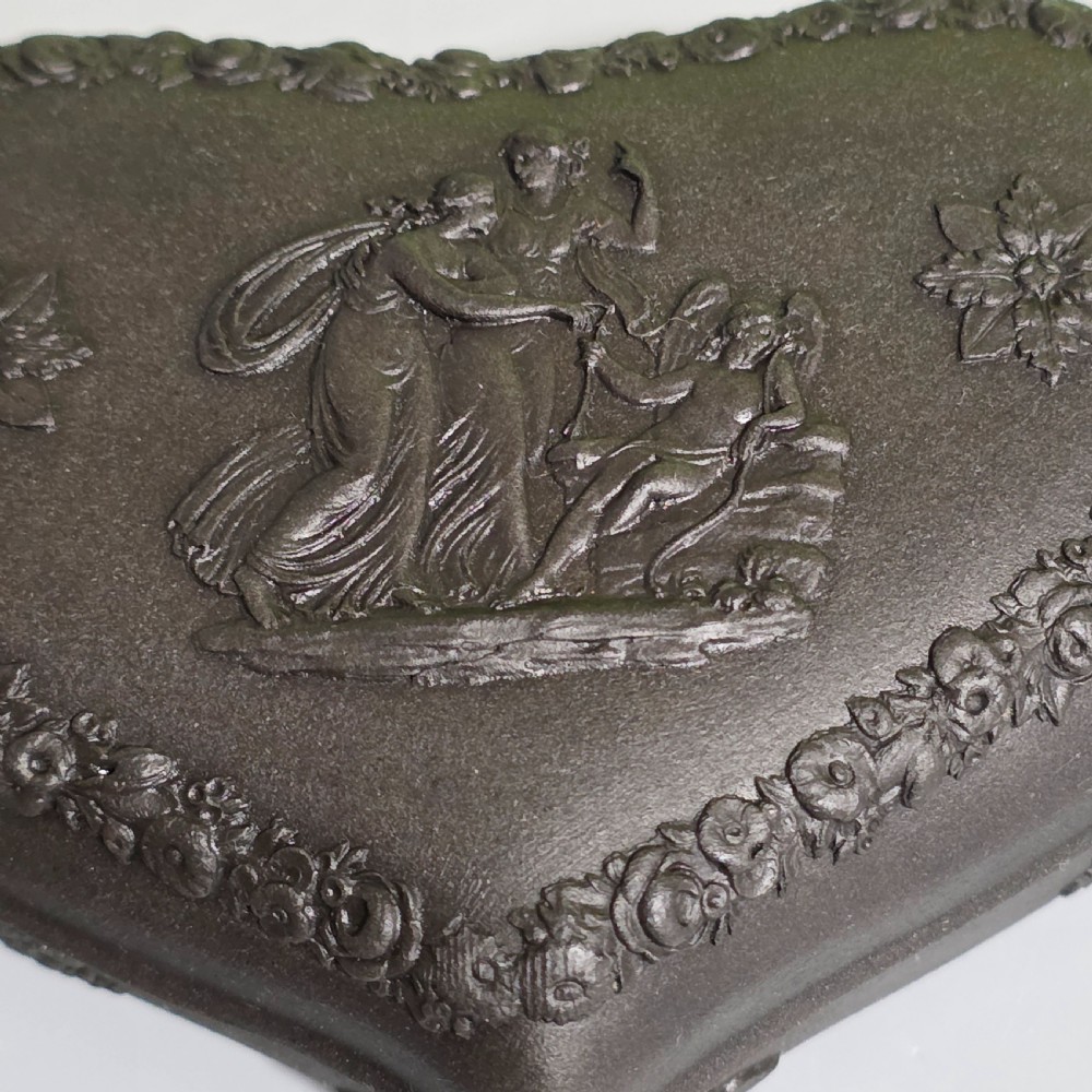 A kidney-shaped matte black basalt stoneware trinket box featuring Neoclassical relief figures, rosettes, and floral garland borders. View II
