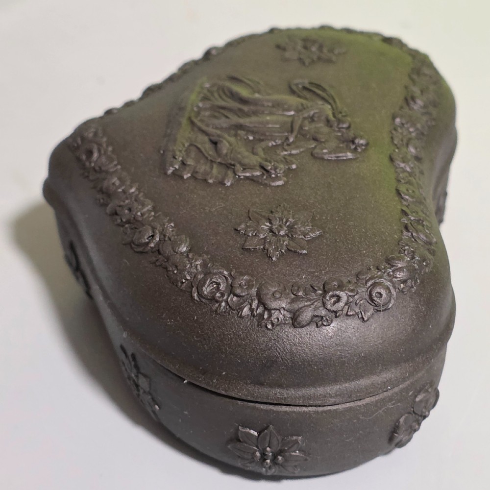 A kidney-shaped matte black basalt stoneware trinket box featuring Neoclassical relief figures, rosettes, and floral garland borders. View III