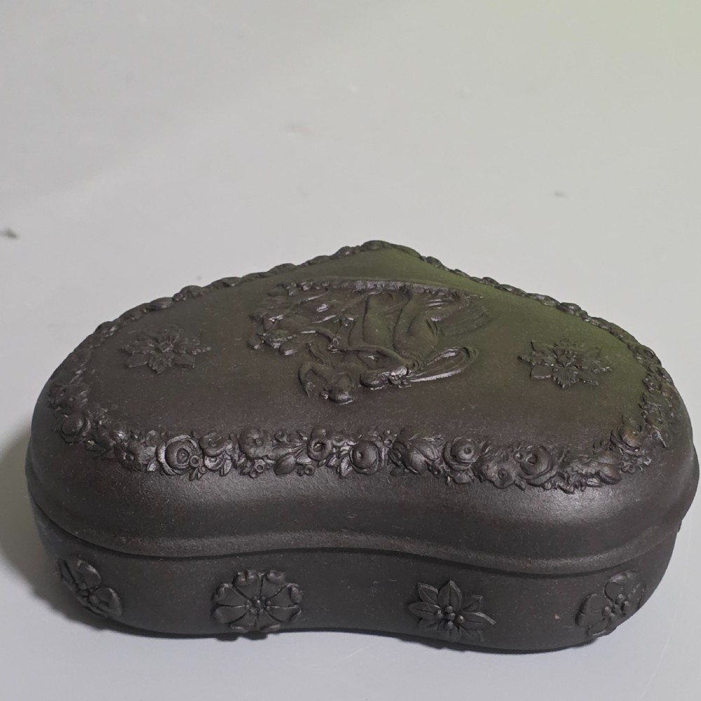 A kidney-shaped matte black basalt stoneware trinket box featuring Neoclassical relief figures, rosettes, and floral garland borders. View IV
