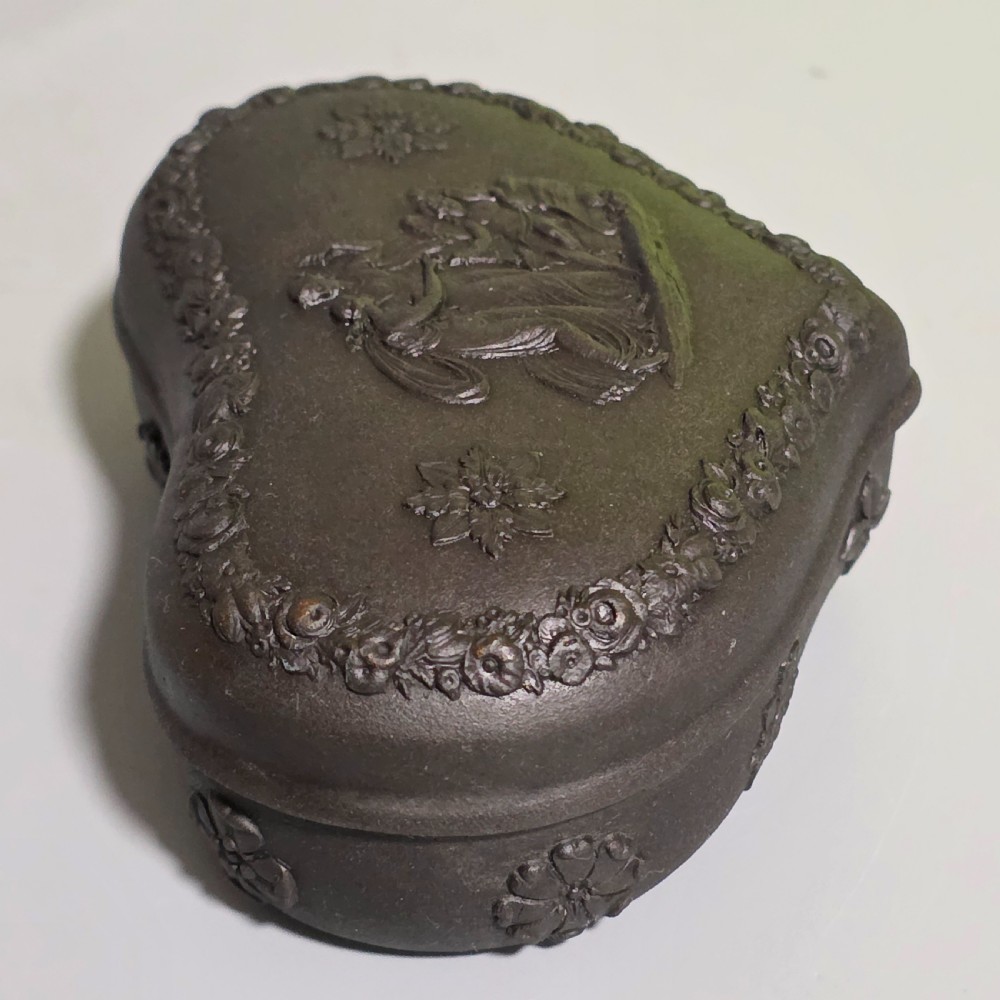 A kidney-shaped matte black basalt stoneware trinket box featuring Neoclassical relief figures, rosettes, and floral garland borders. View V