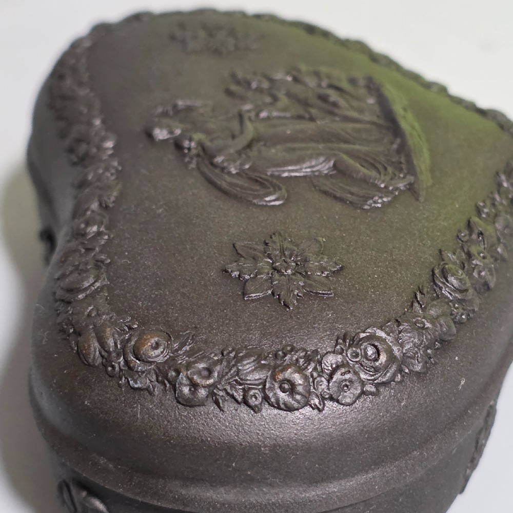 A kidney-shaped matte black basalt stoneware trinket box featuring Neoclassical relief figures, rosettes, and floral garland borders. View VI