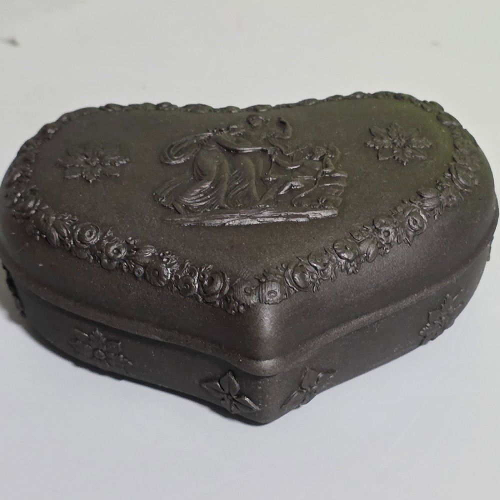 A kidney-shaped matte black basalt stoneware trinket box featuring Neoclassical relief figures, rosettes, and floral garland borders. View VII