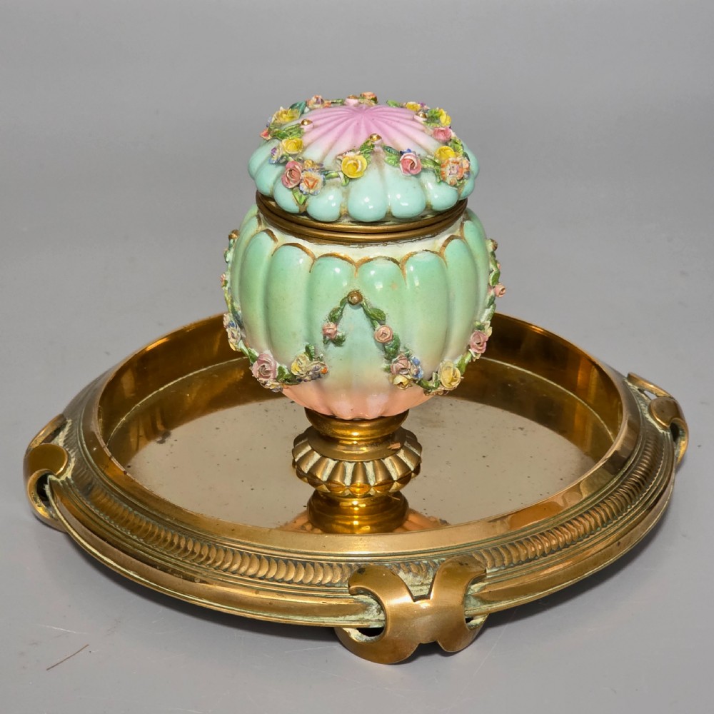 A fluted pastel green and pink porcelain inkwell decorated with applied floral swags, mounted on a turned brass pedestal within a circular brass tray base.