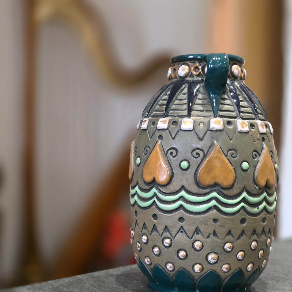 An Art Deco ceramic amphora vase featuring two green handles, raised geometric patterns, heart-shaped motifs, and a glazed teal, grey, and ochre colour scheme. View II