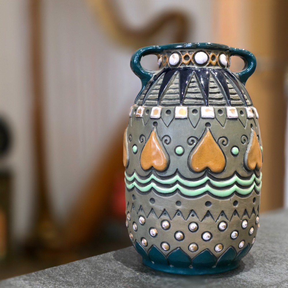 An Art Deco ceramic amphora vase featuring two green handles, raised geometric patterns, heart-shaped motifs, and a glazed teal, grey, and ochre colour scheme.