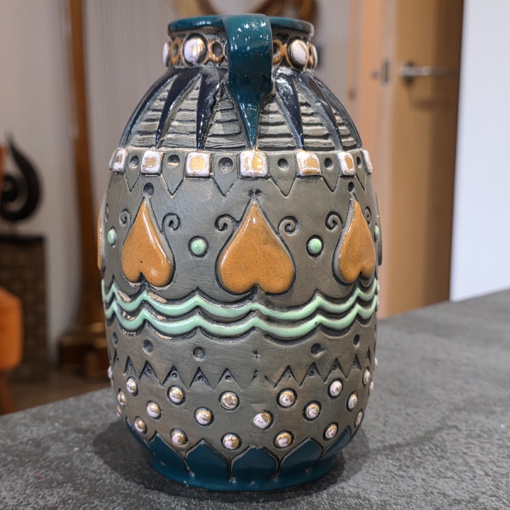 An Art Deco ceramic amphora vase featuring two green handles, raised geometric patterns, heart-shaped motifs, and a glazed teal, grey, and ochre colour scheme. View V