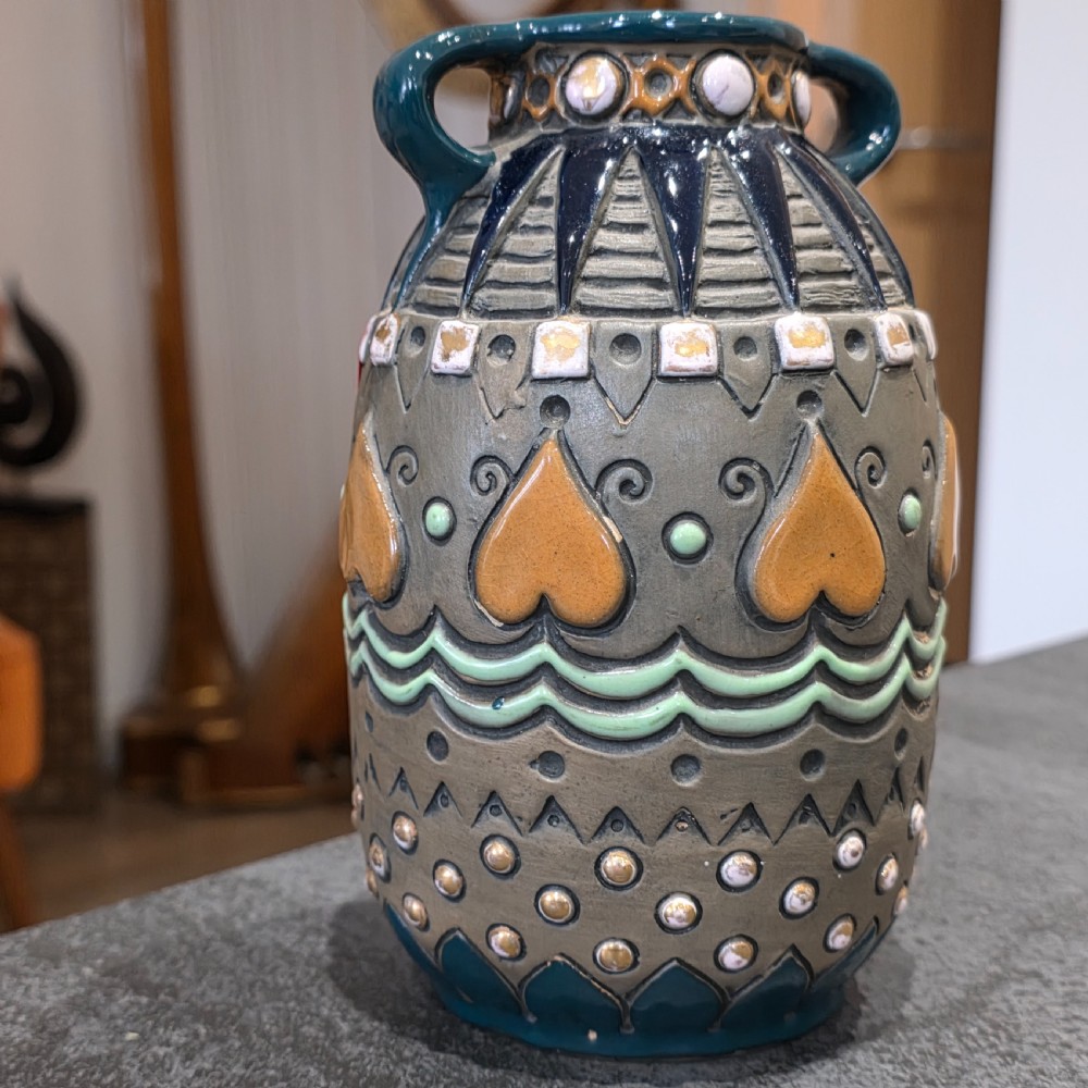 An Art Deco ceramic amphora vase featuring two green handles, raised geometric patterns, heart-shaped motifs, and a glazed teal, grey, and ochre colour scheme. View VI