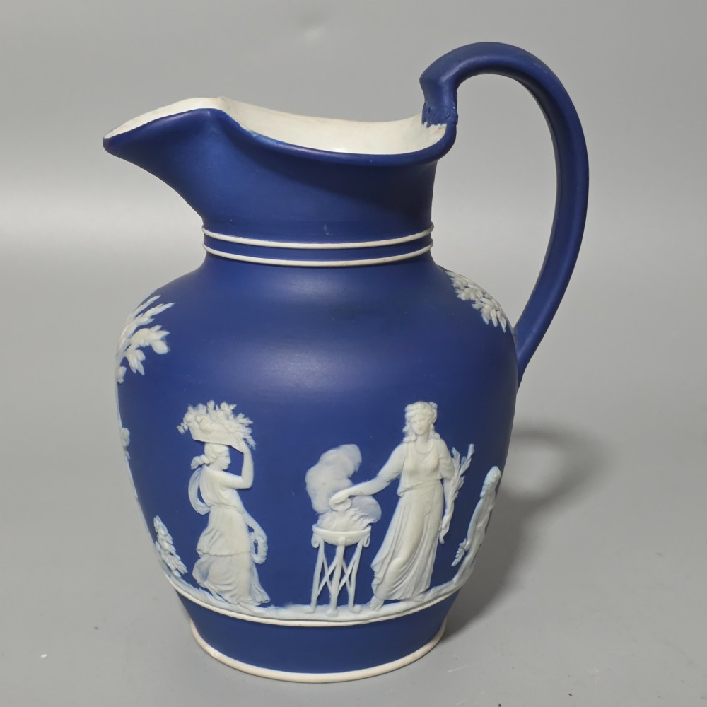 A cobalt blue stoneware jug featuring white raised neoclassical figures in relief with a high arched handle and curved spout.