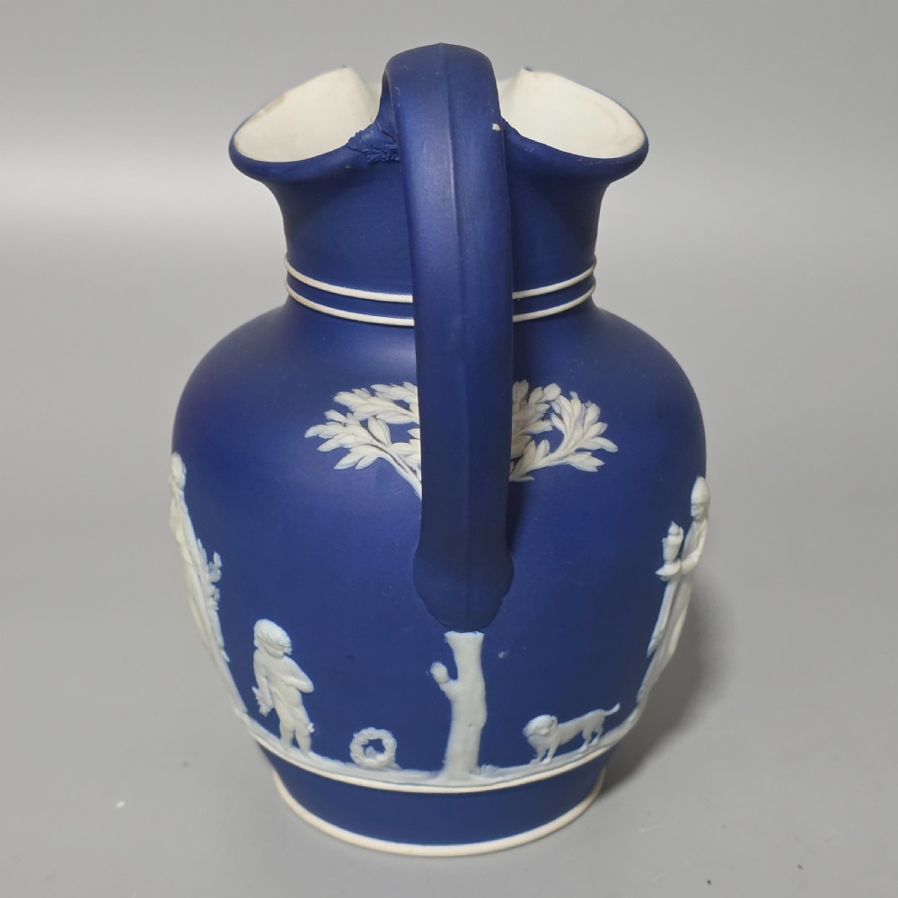 A cobalt blue stoneware jug featuring white raised neoclassical figures in relief with a high arched handle and curved spout. View II