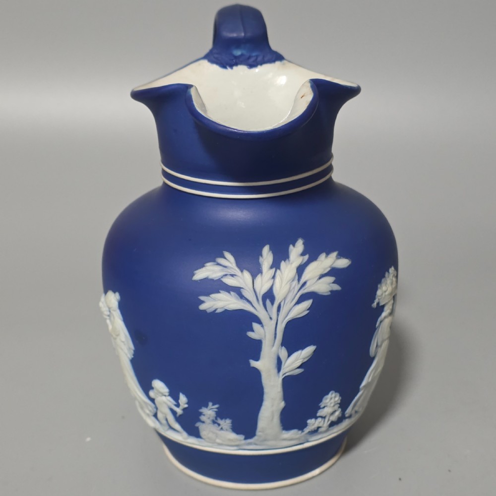 A cobalt blue stoneware jug featuring white raised neoclassical figures in relief with a high arched handle and curved spout. View IV