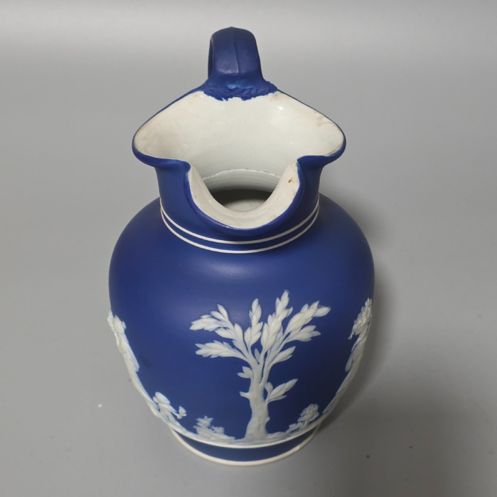 A cobalt blue stoneware jug featuring white raised neoclassical figures in relief with a high arched handle and curved spout. View V