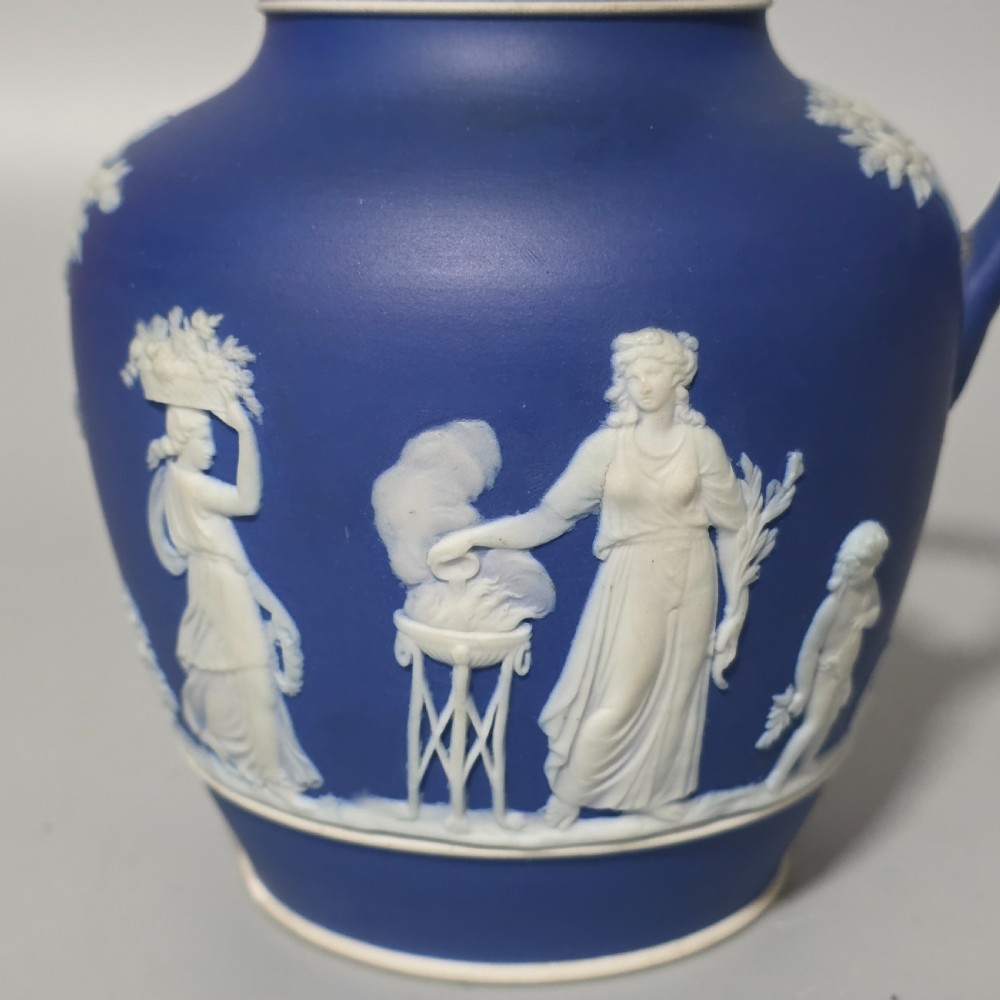 A cobalt blue stoneware jug featuring white raised neoclassical figures in relief with a high arched handle and curved spout. View VII