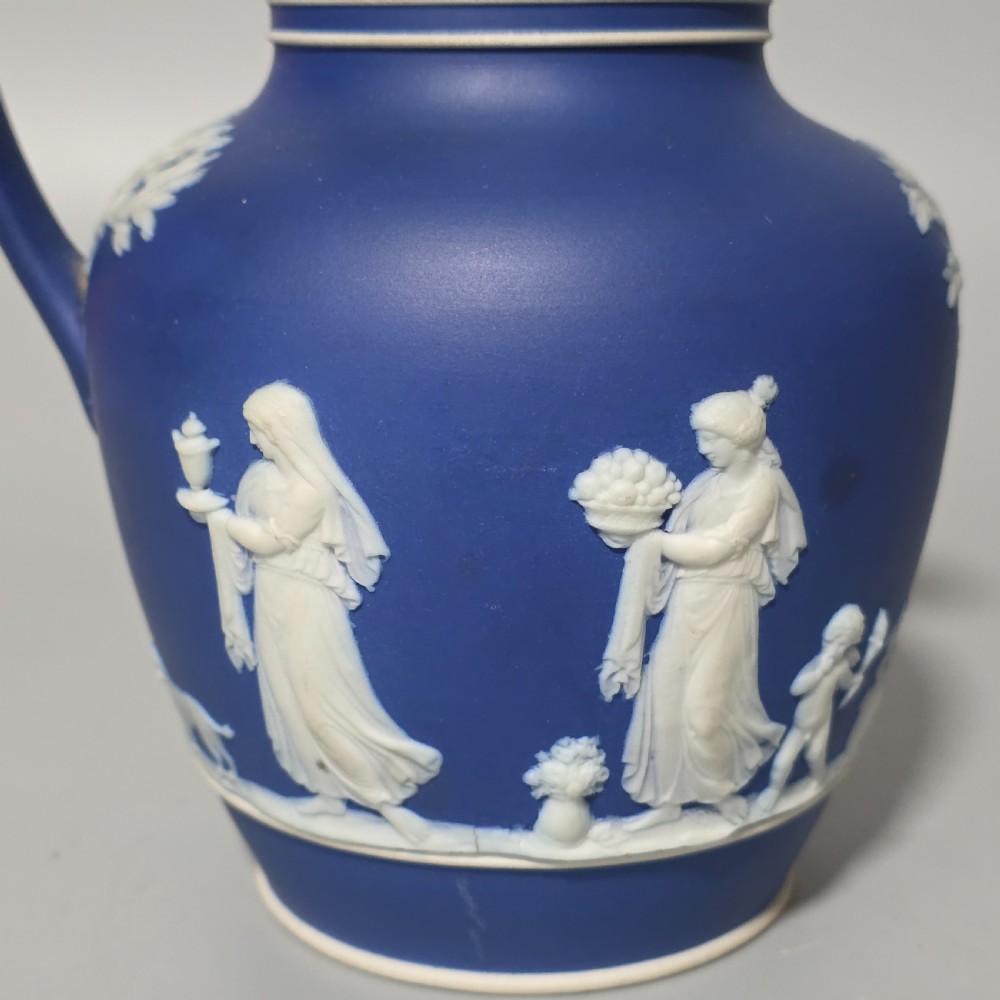 A cobalt blue stoneware jug featuring white raised neoclassical figures in relief with a high arched handle and curved spout. View VIII
