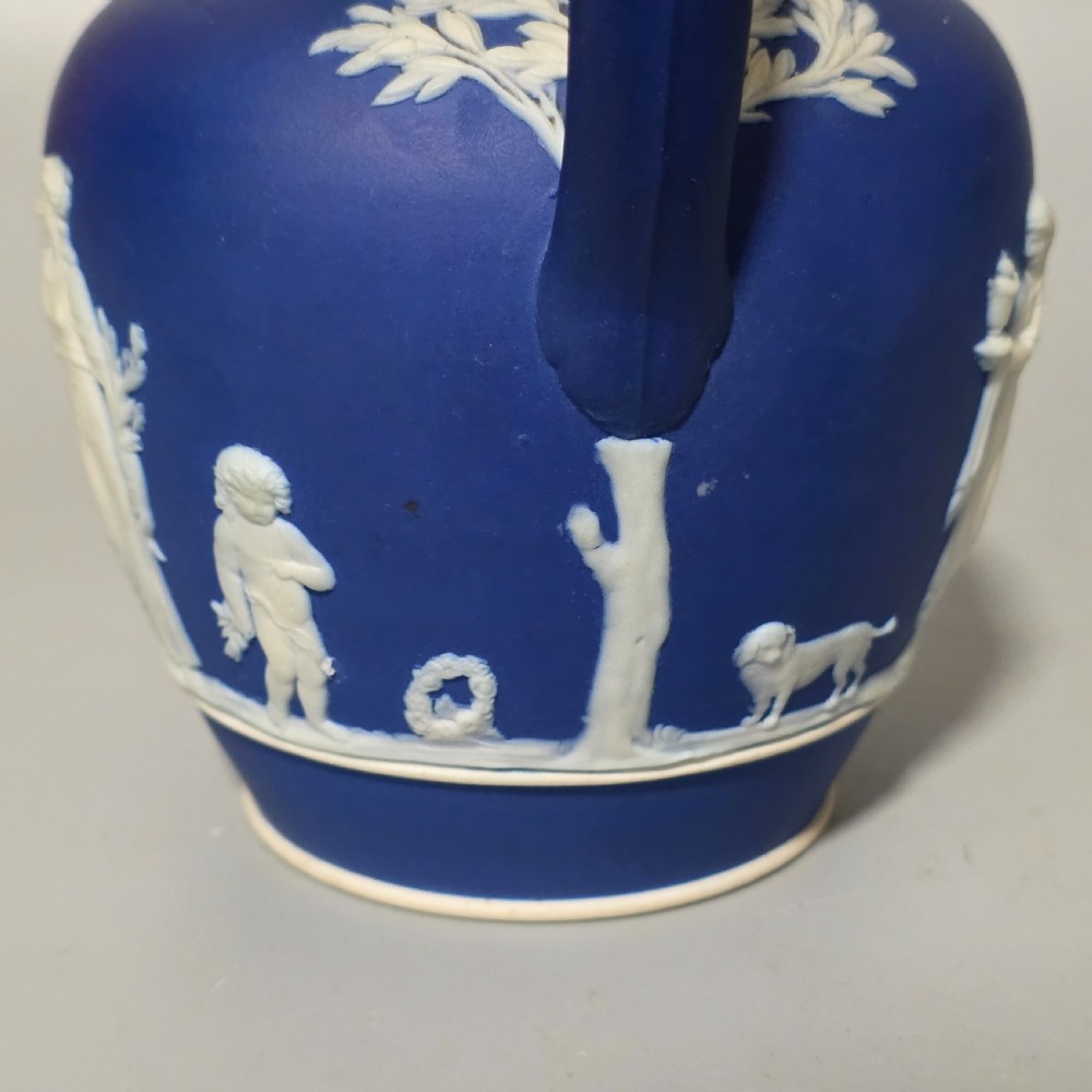 A cobalt blue stoneware jug featuring white raised neoclassical figures in relief with a high arched handle and curved spout. View X