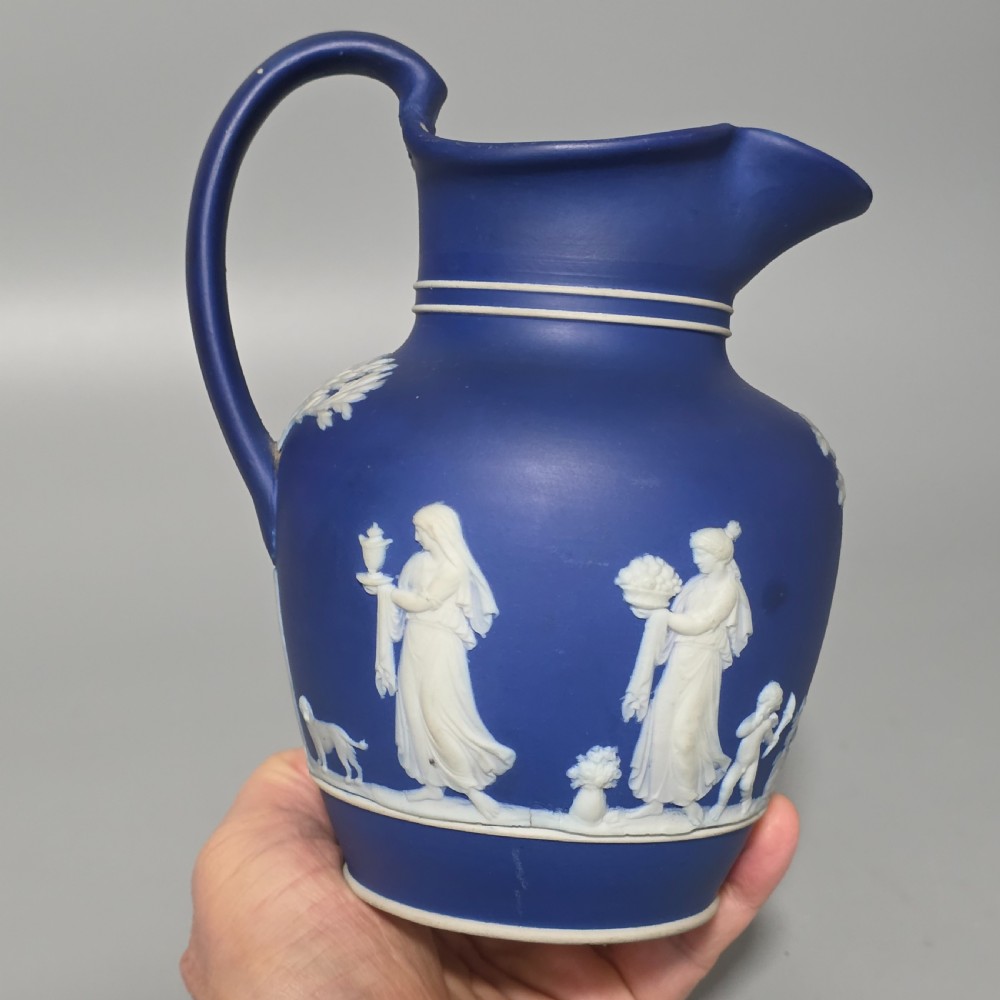 A cobalt blue stoneware jug featuring white raised neoclassical figures in relief with a high arched handle and curved spout. View XIV
