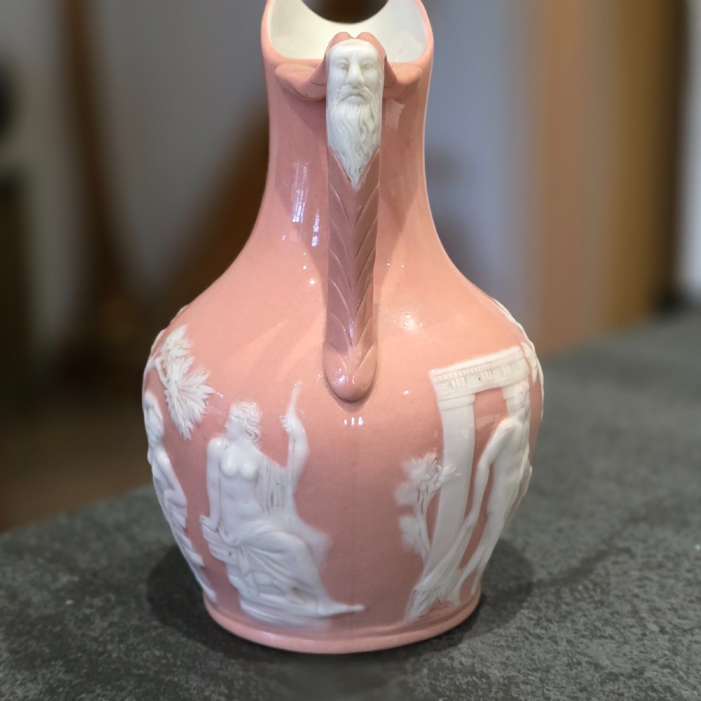A mid-to-late 19th-century salmon pink glazed porcelain pitcher featuring white low-relief classical mythological figures and a sculpted mask handle.