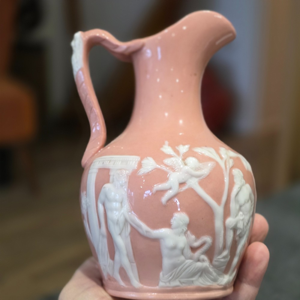 A mid-to-late 19th-century salmon pink glazed porcelain pitcher featuring white low-relief classical mythological figures and a sculpted mask handle. View V