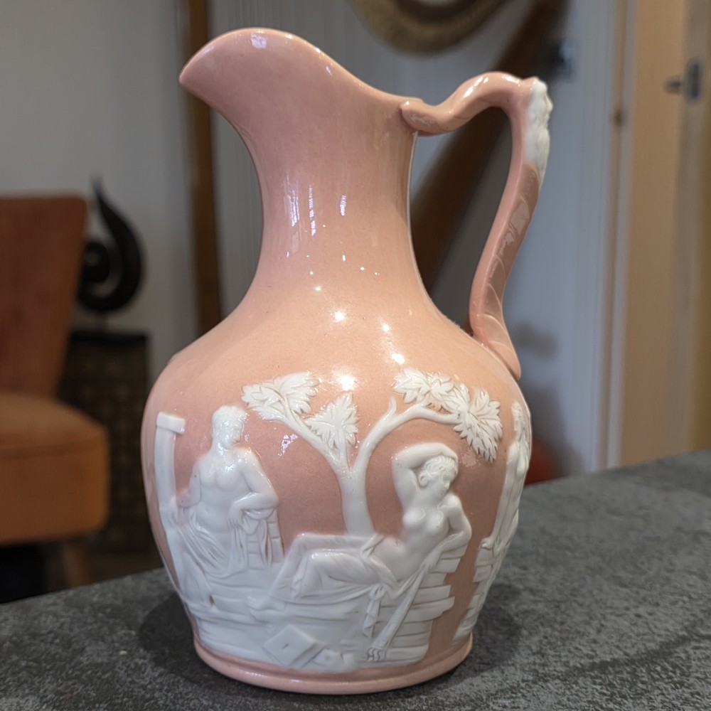 A mid-to-late 19th-century salmon pink glazed porcelain pitcher featuring white low-relief classical mythological figures and a sculpted mask handle. View VI