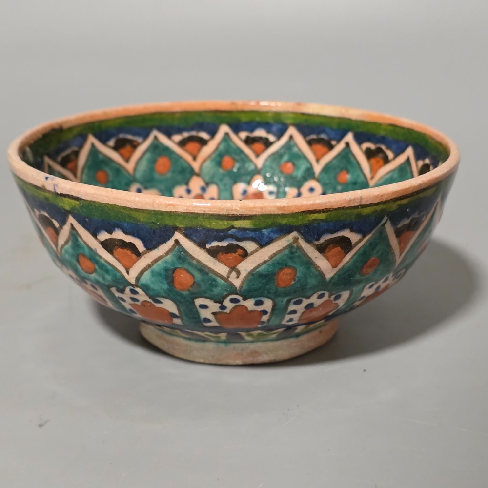 A circular hand-painted antique Middle Eastern ceramic bowl decorated with turquoise, cobalt blue, and terracotta geometric motifs.
