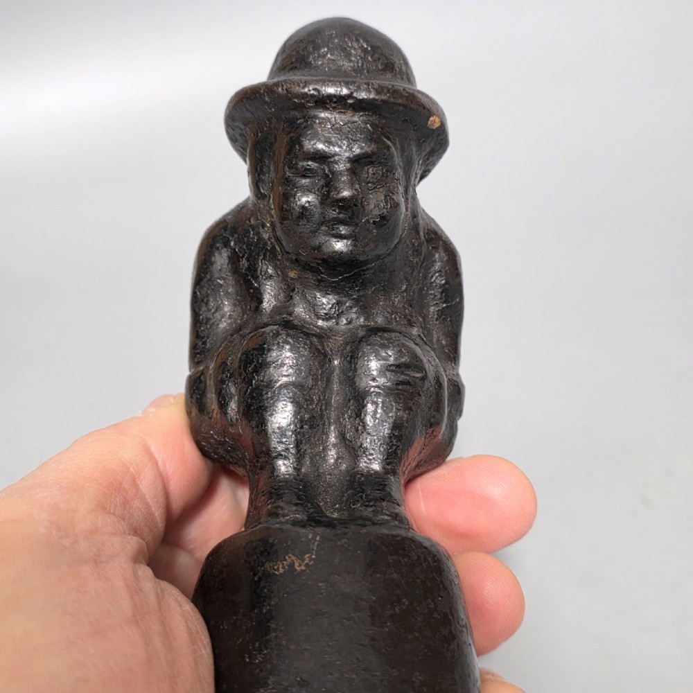 A nineteenth-century dark cast iron paperweight shaped as a crouching male figure in a hat set upon a cylindrical pedestal. View II