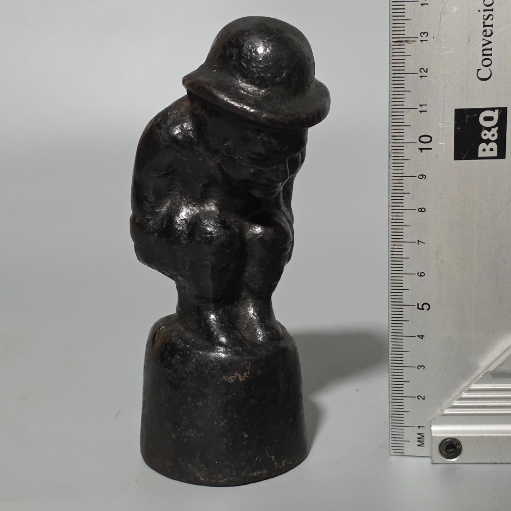 A nineteenth-century dark cast iron paperweight shaped as a crouching male figure in a hat set upon a cylindrical pedestal. View X