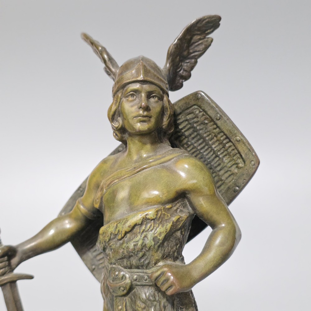 A patinated bronze sculpture depicting the mythical hero Siegfried standing victorious with a sword, winged helmet, and shield above a dragon base. View X
