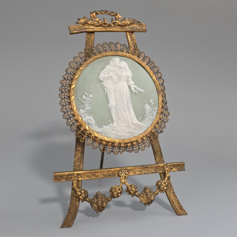 A pale green and white bisque porcelain oval plaque depicting two figures in high relief, supported on an ornate gilt metal easel.